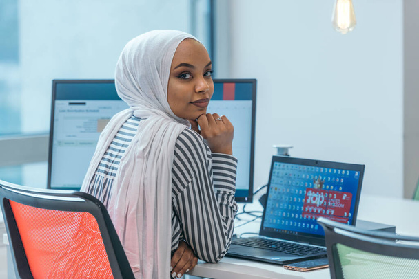 Üzleti oktatás és irodai koncepció. Afrikai-amerikai üzletasszony, hidzsábot visel, dolgozik, számítógépen gépel. Női vállalkozó ül az asztalán. - Fotó, kép