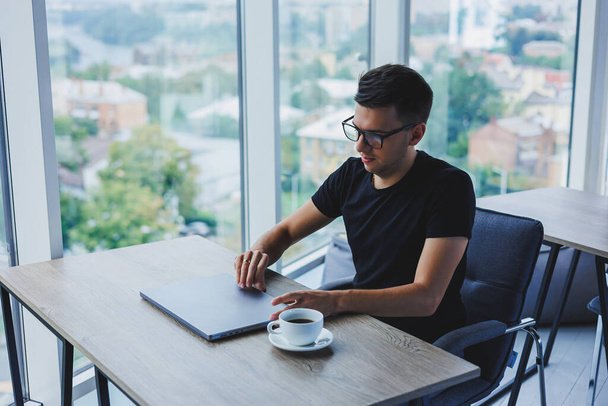 Веселый бизнесмен в очках сидит за своим столом, работая с ноутбуком в офисе. Бизнесмен сидит и работает за ноутбуком и пьет кофе. Начало дня в офисе - Фото, изображение