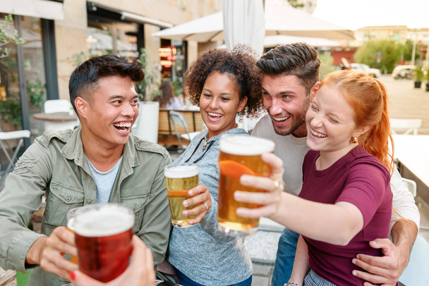 Group of happy diverse friends clinking glasses with beer while spending time together on terrace of outdoor cafe in city - Foto, Bild
