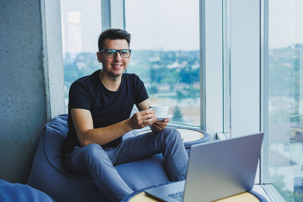 Smiling businessman looking for something and typing on a laptop and drinking fresh black coffee. The concept of a modern successful person. Young focused guy with glasses in an open office - Zdjęcie, obraz