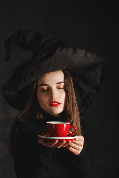 Чарівна молода дівчина в капелюсі відьми з чашкою гарячої кави в руках
 - Фото, зображення