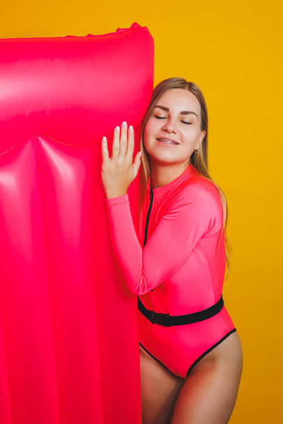 Young slender woman in a pink swimsuit on a yellow background. Girl with an inflatable mattress for swimming in the pool. Summer vacation. - Foto, afbeelding