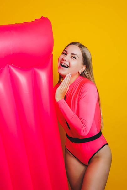 Young slender woman in a pink swimsuit on a yellow background. Girl with an inflatable mattress for swimming in the pool. Summer vacation. - Fotografie, Obrázek