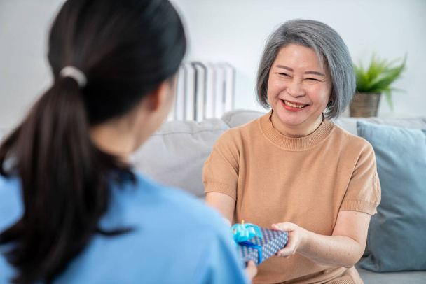 A young caregiver hand over to her senior patient a blue gift box with blue ribbons at a contented living room. - Foto, immagini
