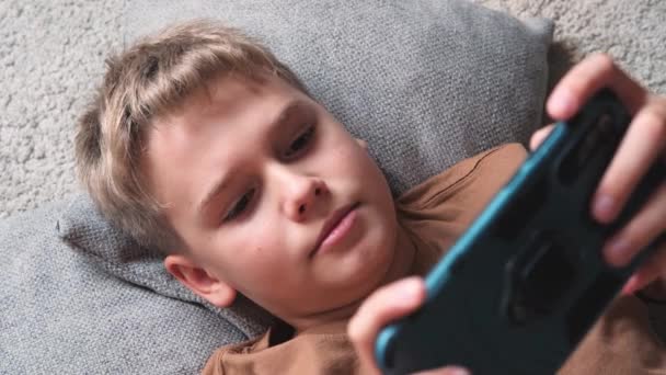 Pre teenager uses smartphone phone while lying on the floor at home. Close-up face of a boy playing games on the phone - Séquence, vidéo