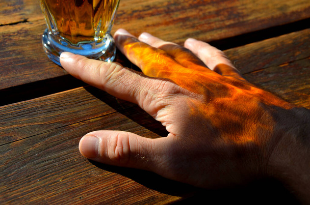 Золотой мед пива светит через стекло на руку человека, ладонь свободно покоится на деревянном верху стола. мужчина расслабляется с любимым напитком и делает рекламу - Фото, изображение