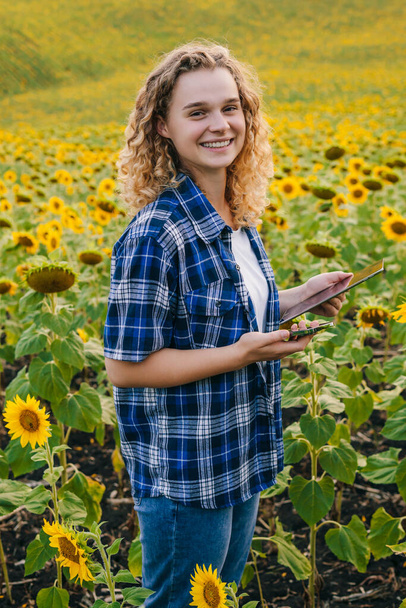 Πορτρέτο ενός χαμογελαστού σγουρού κοριτσιού που εργάζεται στον τομέα του ηλίανθου κάνει την ανάλυση της ανάπτυξης της καλλιέργειας των φυτών. Έξυπνη γεωργία και γεωργία ακριβείας. Ηλίανθος - Φωτογραφία, εικόνα