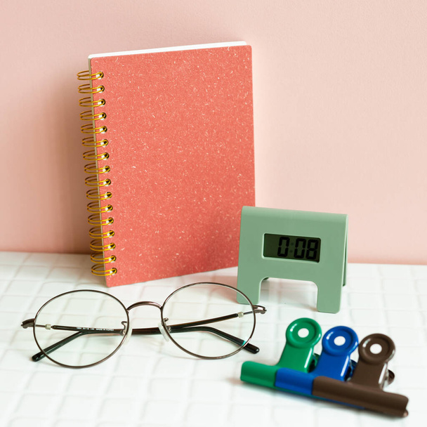 Office desk table with notebook, eyeglasses, clock, clip, supplies on white desk. pink wall background - 写真・画像