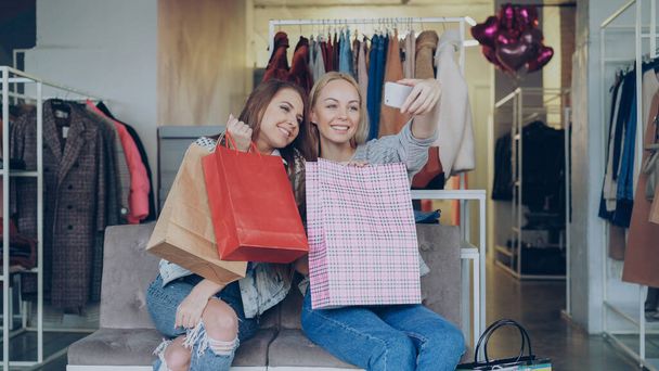 Amigas atraentes estão usando o smartphone para fazer selfie enquanto estão sentadas na loja de roupas femininas com muitos sacos de papel coloridos. Eles estão sorrindo, posando, rindo, gesticulando
. - Foto, Imagem