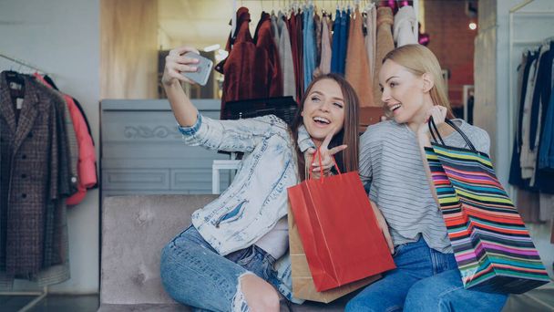 Cute girls are sitting and making selfie with colourful paper bags using smartphone in luxurious womens clothing boutique, then watching pictures together. They are smiling and laughing carelessly. - Foto, imagen