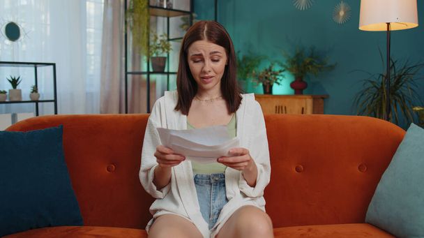 Stressed girl at home room looking at unpaid bank debt bills, doing paperwork, planning budget, calculate finances mortgage payments. Displeased young woman with paper bills sitting on orange couch - Zdjęcie, obraz