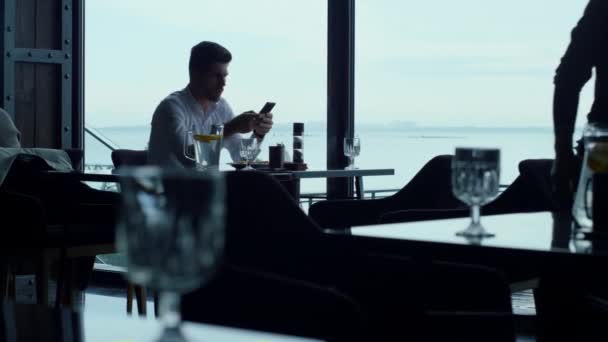 Successful entrepreneur working on smartphone at breakfast time in stylish sky bar. Busy employee sending emails at lunch break in restaurant. Unrecognizable male silhouette walking hotel cafe hall. - Záběry, video