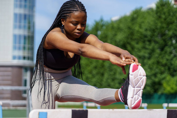Young fit black woman enjoys sports activity at playground of big residential complex on sunny summer day. African American lady puts leg on metal hurdles barrier to stretch before intense training - Photo, Image