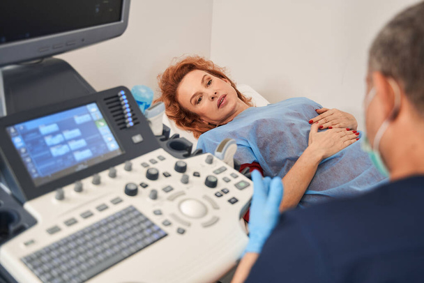 Cropped view of the serious caucasian patient woman talking with her mature doctor during the ultrasound therapy at the hospital - Фото, изображение