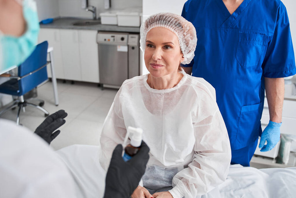 Happy woman sitting at the hospital bed and preparing to the gastroscopy. Doctor and nurse at the background. Stock photo - Photo, image