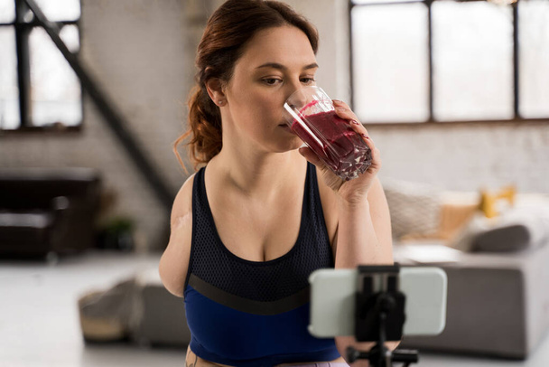 Healthy lifestyle. Portrait view of the woman without hand drinking smoothie from the berries and looking at the smartphone at the tripod - Фото, изображение