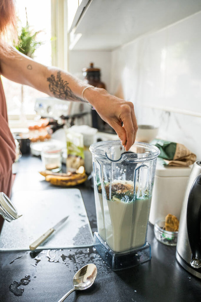 Cropped view of the tattooed woman standing near the table and preparing smoothie for the breakfast while cooking at the kitchen - Foto, Imagen