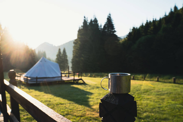 closeup front view of metallic coffee cup in the morning with glamping tent camping ground in the background in a green mountain meadow surrounded by fir tree forest - Foto, Imagem