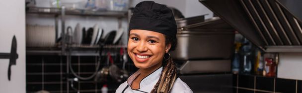 happy and young african american chef in hat smiling in professional kitchen, banner - Φωτογραφία, εικόνα