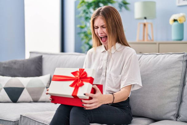 Beautiful woman holding gift angry and mad screaming frustrated and furious, shouting with anger. rage and aggressive concept.  - Fotoğraf, Görsel