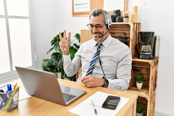 Latynoski biznesmen w średnim wieku, pracujący w biurze, noszący słuchawki operatora, pokazujący i wskazujący palcami numer trzy, uśmiechający się pewny siebie i szczęśliwy..  - Zdjęcie, obraz