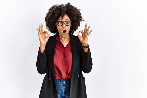 Afro-Amerikaanse vrouw met afro-haar dragen business jas en bril kijken verrast en geschokt doen ok goedkeuring symbool met vingers. gekke uitdrukking  - Foto, afbeelding
