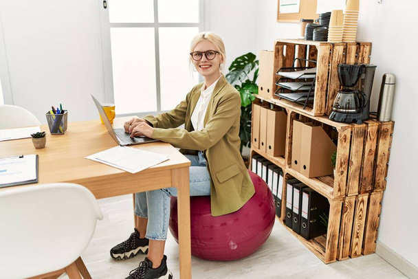 Piękny biały biznes kobieta pracuje w biurze siedzi na pilates ball patrząc pozytywnie i szczęśliwy stojąc i uśmiechając się z pewnym uśmiechem pokazując zęby  - Zdjęcie, obraz