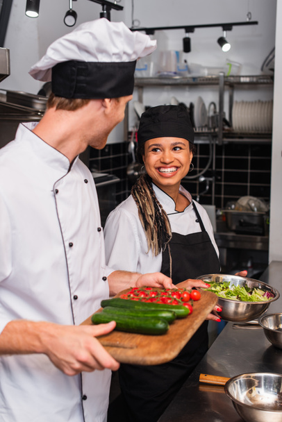 chef and cheerful african american sous chef standing with ingredients in kitchen  - Photo, Image