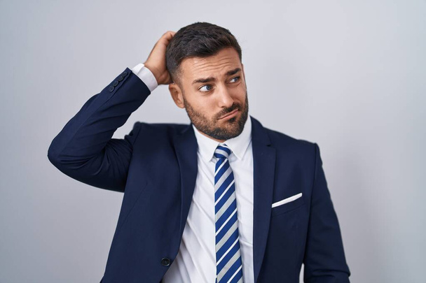 Handsome hispanic man wearing suit and tie confuse and wondering about question. uncertain with doubt, thinking with hand on head. pensive concept.  - Фото, изображение
