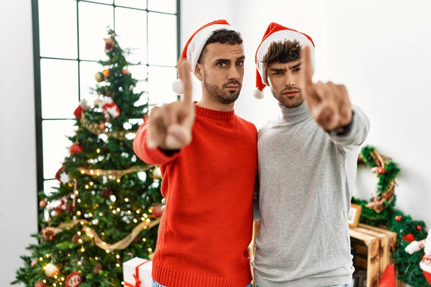 Young gay couple standing by christmas tree wearing hat pointing with finger up and angry expression, showing no gesture  - Photo, Image