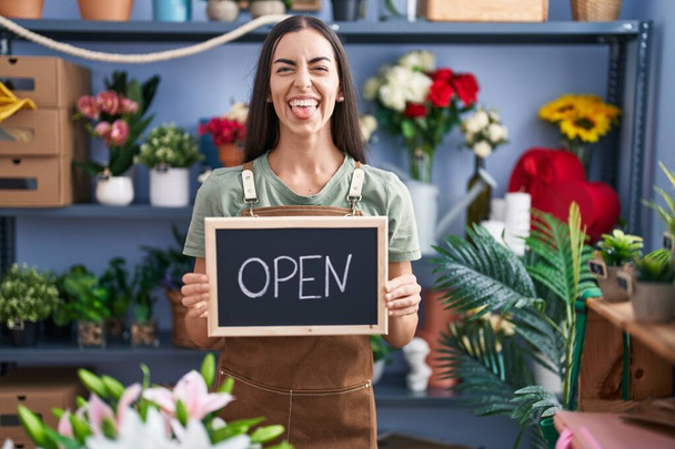 Jeune femme brune travaillant chez le fleuriste tenant signe ouvert langue collante heureux avec une expression drôle.  - Photo, image