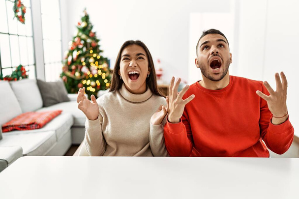Giovane coppia ispanica seduta sul tavolo da albero di Natale pazzo e pazzo urlando e urlando con espressione aggressiva e braccia alzate. concetto di frustrazione.  - Foto, immagini