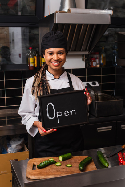 happy african american chef holding chalkboard with open lettering in kitchen - Φωτογραφία, εικόνα
