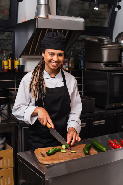 cheerful african american woman in apron cutting cucumber in modern kitchen  - Foto, Imagen