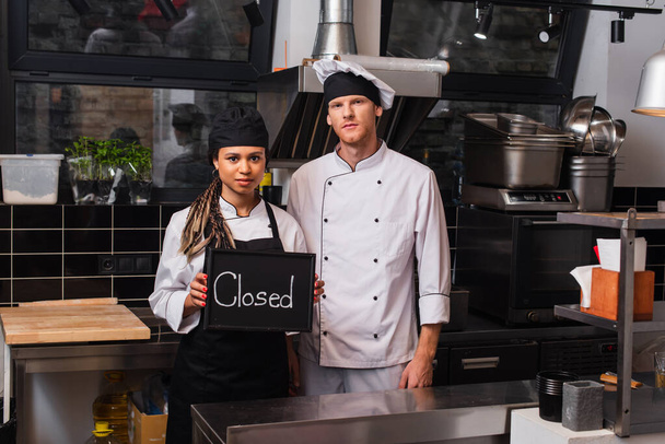 young african american woman holding chalkboard with closed lettering near chef in kitchen - Φωτογραφία, εικόνα