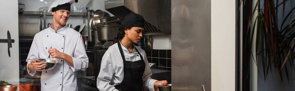 cheerful chef whisking eggs and looking at african american colleague in kitchen, banner - Фото, изображение
