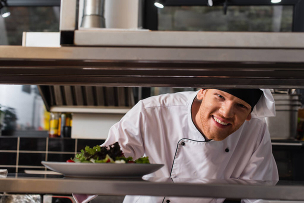happy chef holding plate with freshly cooked salad in professional kitchen - Photo, Image