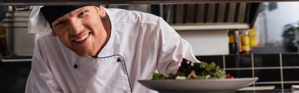 happy chef holding plate with freshly cooked salad in professional kitchen, banner - Foto, Imagem