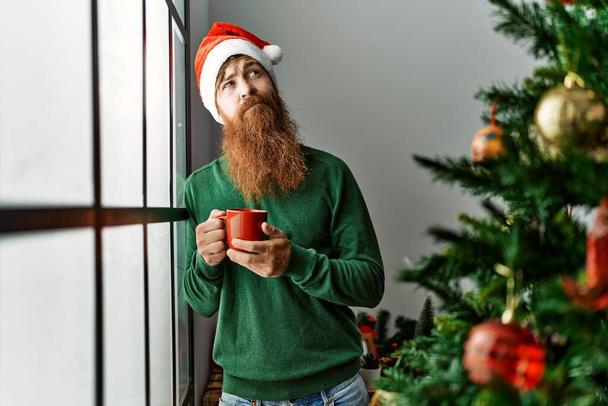 Молодой рыжий мужчина пьет кофе, стоя у рождественской елки дома - Фото, изображение