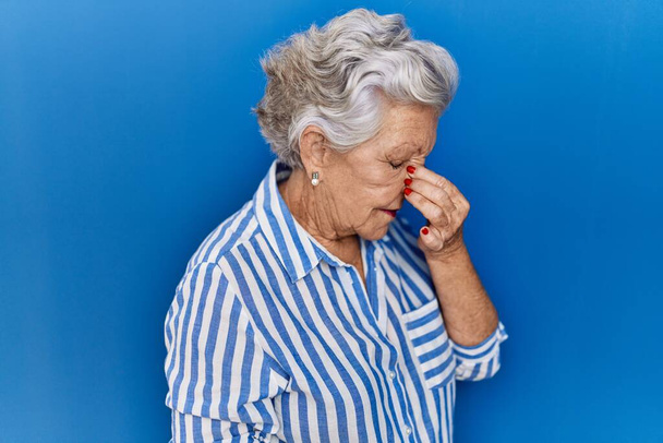 Ηλικιωμένη γυναίκα με γκρίζα μαλλιά στέκεται πάνω από το μπλε φόντο κουρασμένος τρίψιμο μύτη και τα μάτια αίσθημα κόπωσης και πονοκέφαλο. άγχος και απογοήτευση έννοια.  - Φωτογραφία, εικόνα