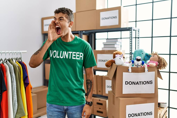 Młody, przystojny Latynos noszący koszulę wolontariusza na darowiznach krzyczy i krzyczy głośno, by stanąć z ręką w twarz. Koncepcja komunikacji.  - Zdjęcie, obraz