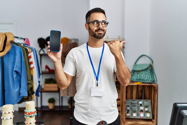 Young hispanic man with beard working as manager at retail boutique holding smartphone pointing thumb up to the side smiling happy with open mouth  - 写真・画像