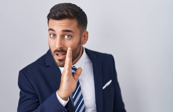 Hombre hispano guapo usando traje y corbata de la mano en la boca contando rumores secretos, susurrando conversación maliciosa  - Foto, imagen