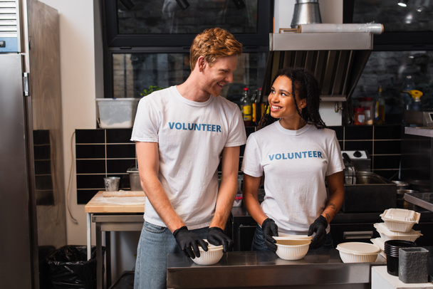 happy multiethnic volunteers in t-shirts preparing plastic containers with food in kitchen  - Foto, imagen