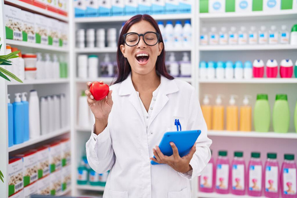 Junge hispanische Frau, die in der Apotheke arbeitet, hält rotes Herz, lächelt und lacht laut, weil lustig verrückter Witz.  - Foto, Bild