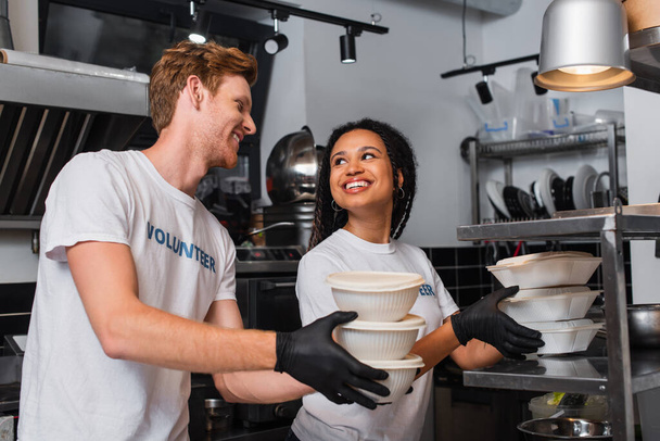 усміхнені багатоетнічні добровольці в футболках, що тримають пластикову тару з їжею на кухні
  - Фото, зображення