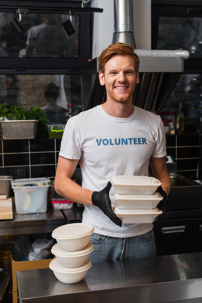 redhead man in t-shirt with volunteer lettering smiling and holding plastic containers in kitchen  - Photo, image