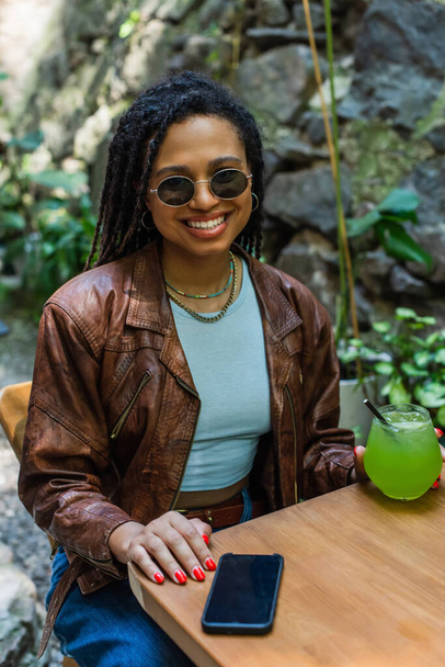 happy african american woman in sunglasses holding glass with green alcohol cocktail in outdoor terrace  - Фото, изображение