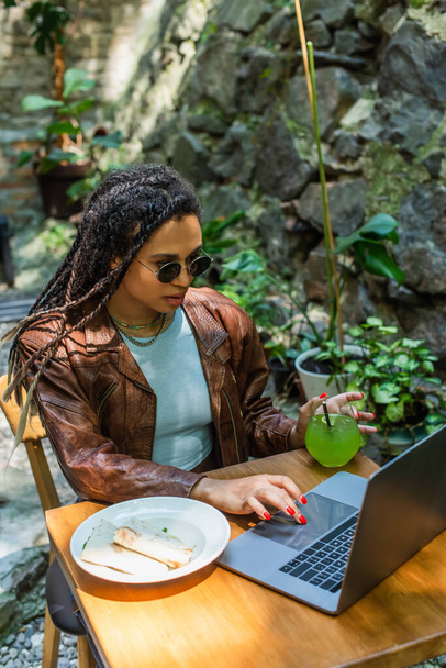 young african american woman in sunglasses using laptop near prepared lunch and glass with drink in outdoor cafe terrace - Foto, Imagen