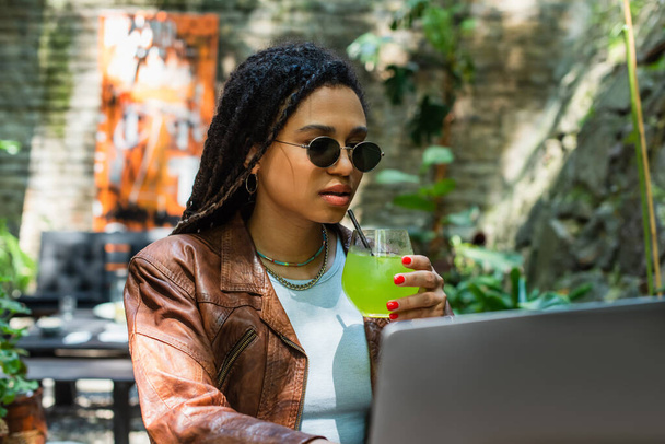 young african american freelancer in sunglasses using laptop and holding glass with cocktail in outdoor terrace - Valokuva, kuva